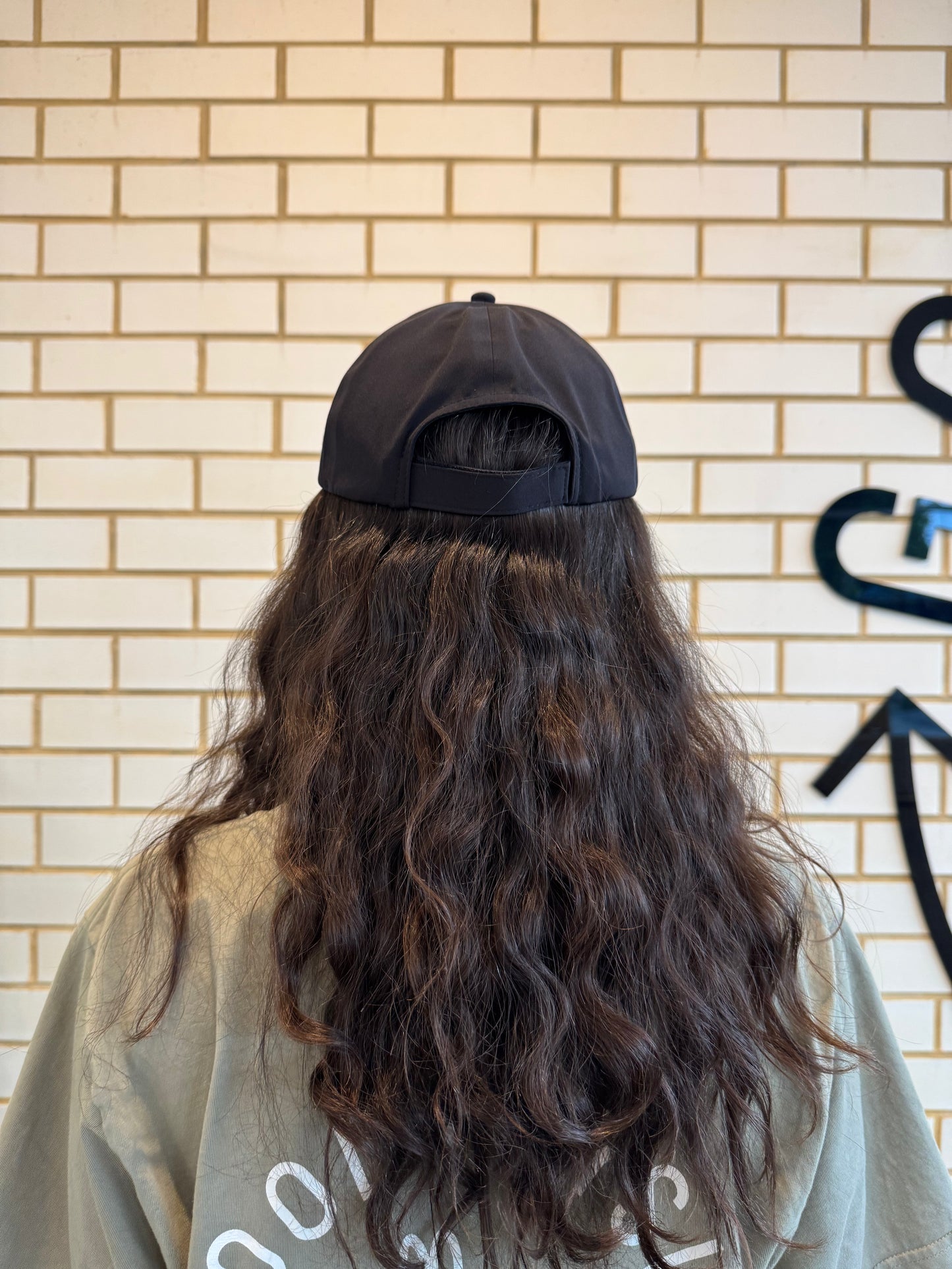 Person with long hair wearing a black cap against a brick wall, showing the back of the cap.