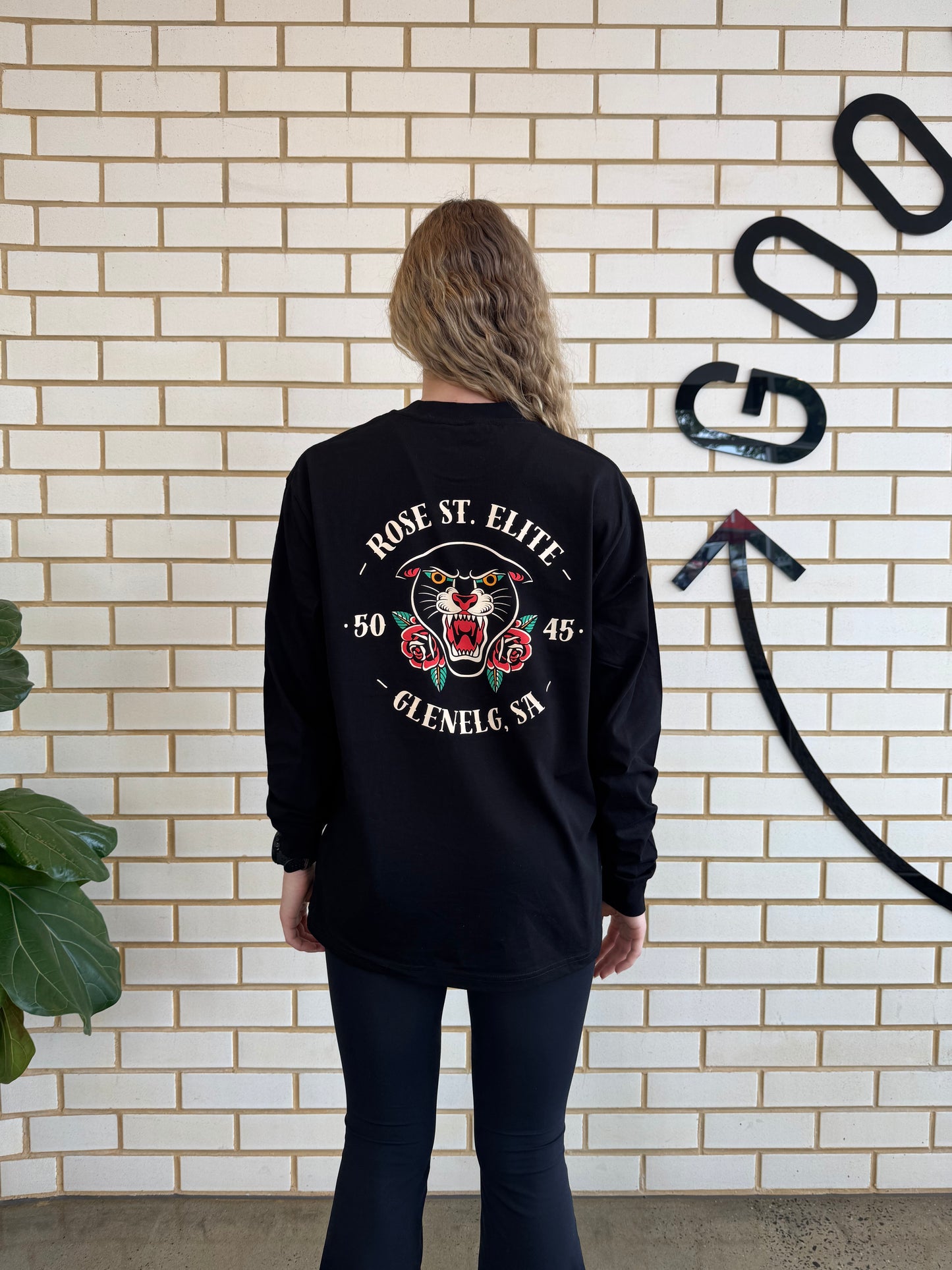 Lady wearing long sleeve black shirt with good physio logo against brick wall.
