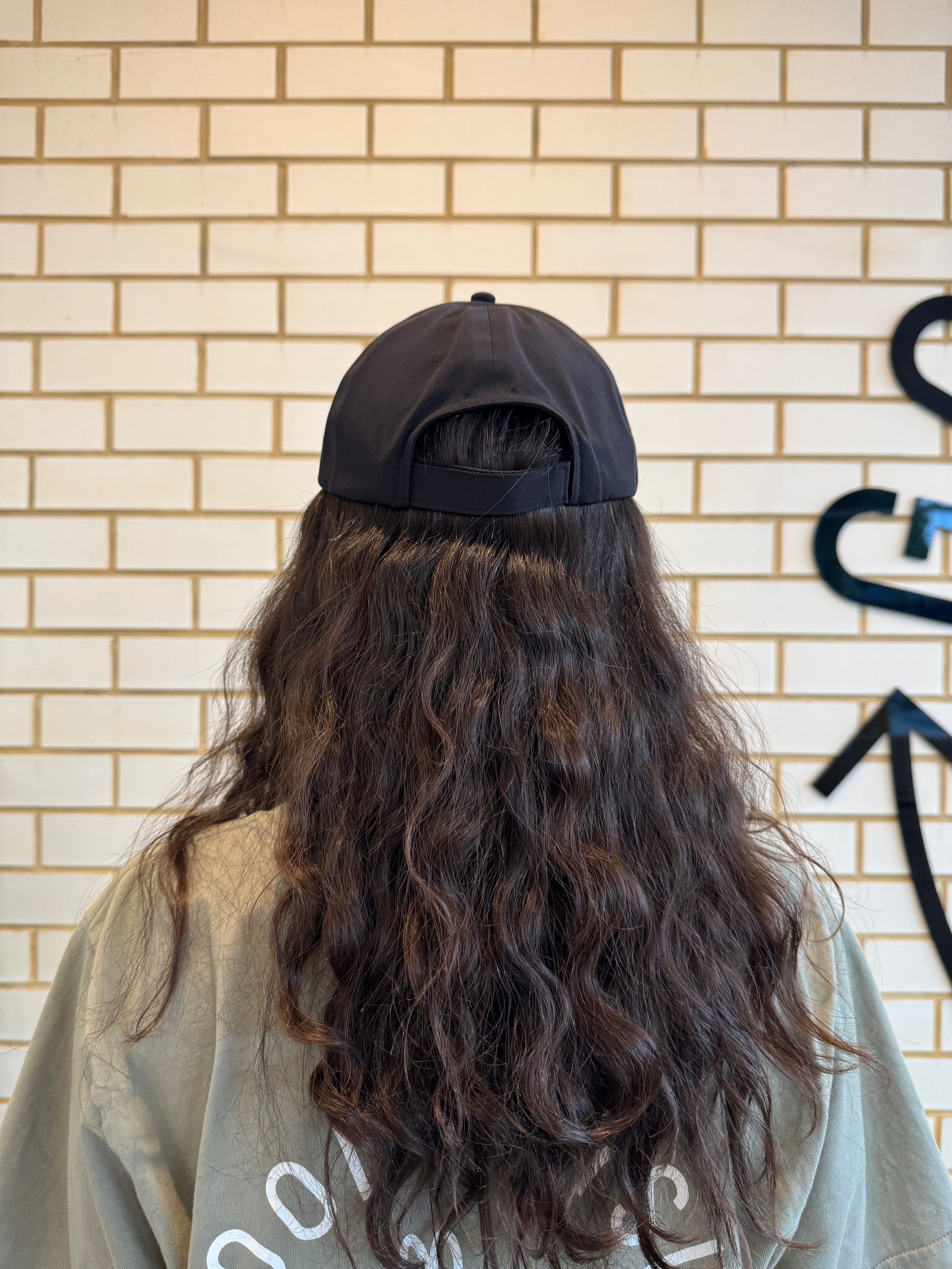 Person with long hair wearing a black cap against a brick wall, showing the back of the cap.