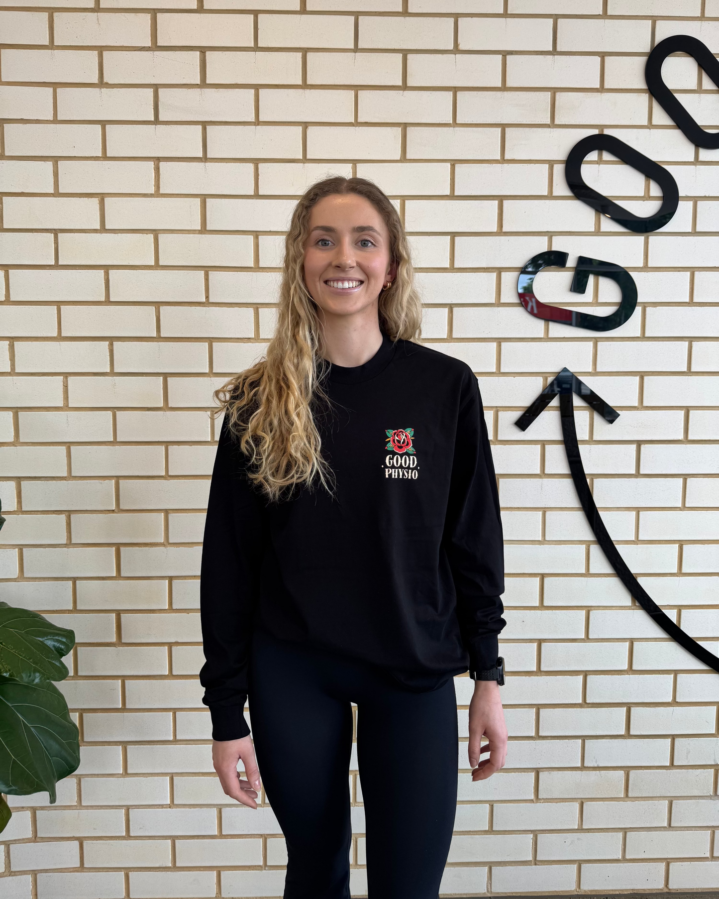Lady wearing a black long sleeve with a good physio logo against a brick wall.