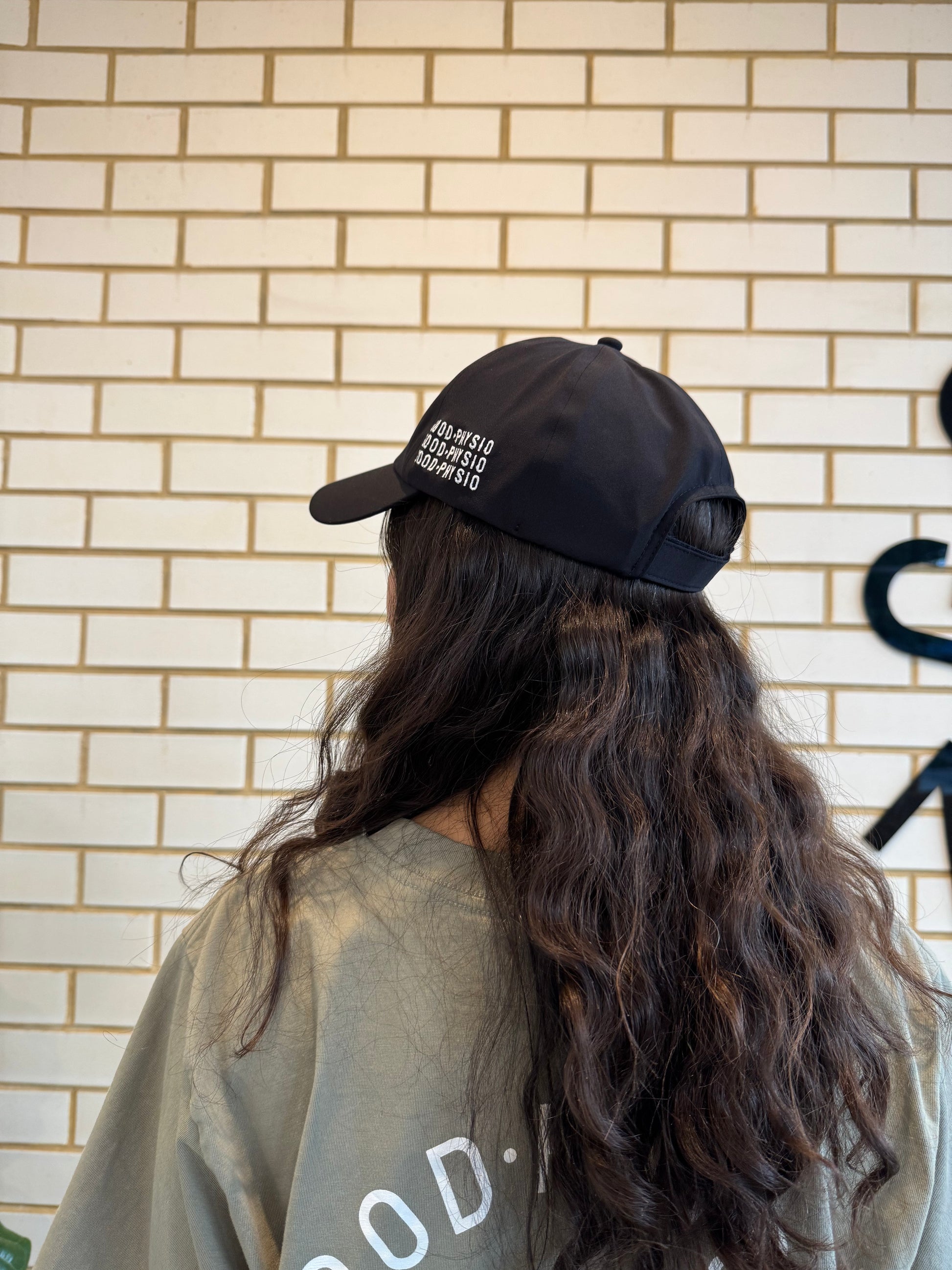 Person wearing a black cap with text against a beige brick wall, showing side of cap.